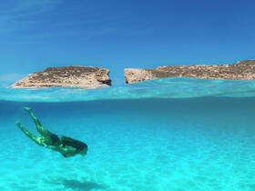 A swimmer explores the vibrant underwater world in crystal-clear waters, showcasing the stunning scenery of rocky formations above the surface.