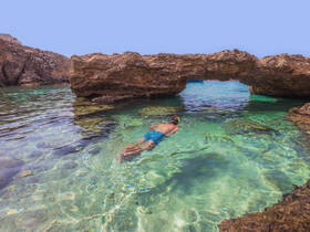 A snorkeler explores the crystal-clear waters beneath a natural rock arch, showcasing the beauty of underwater adventures in stunning coastal scenery.