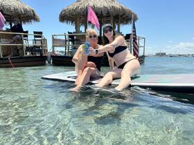 Two friends enjoy a sunny day on a paddleboard, celebrating with drinks in shallow waters — a perfect moment for summer fun and relaxation.