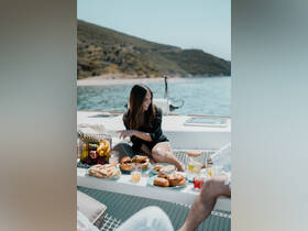 A woman enjoys a delightful spread of snacks and cocktails on a boat, surrounded by fresh fruit — perfect for a relaxing day on the water.