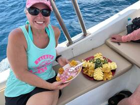 A smiling woman enjoys a delicious spread of fresh fruit and snacks aboard a boat, perfect for a relaxing day on the water.