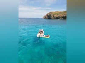 Aerial view of a group enjoying a sunny day on a pontoon boat, with one person relaxing on a floating mat in the crystal-clear waters — a perfect scene for summer adventures.