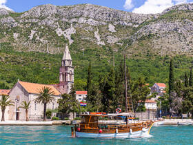 A charming wooden boat navigates the turquoise waters near a picturesque coastal village, with a historic church and lush mountains in the background, perfect for scenic boat tours.