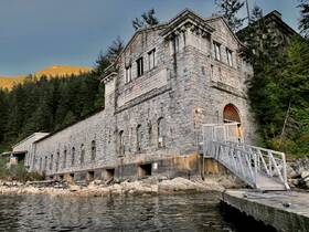 A stone structure near the water, possibly a historical power house, surrounded by forested mountains. The image lacks clarity and detail.