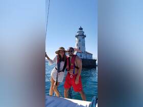 A couple poses joyfully on a boat with a historic lighthouse in the background, capturing a perfect moment of their boating adventure.