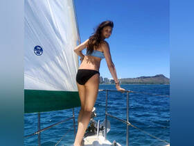 A woman enjoys a sailing adventure, posing on the bow of a boat with stunning ocean views and a landmark in the background, perfect for capturing the essence of a day on the water.