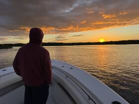 A person stands on the deck of a boat, gazing at a stunning sunset over calm waters, creating a serene atmosphere perfect for evening cruises.