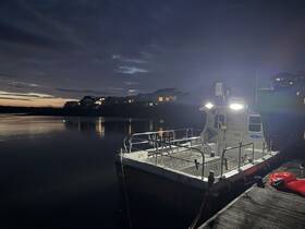 A serene evening view of a docked boat illuminated by lights, reflecting on calm waters as the sun sets behind the houses.