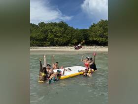 A lively group of friends enjoys a fun day on a floating mat in shallow waters, celebrating with drinks and laughter at the beach.