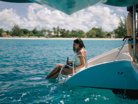 A young woman relaxes on the edge of a boat, dipping her feet into the crystal-clear water, capturing the essence of a perfect beach day.