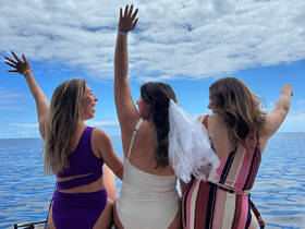 A joyful group of friends celebrates a bachelorette party by the ocean, raising their arms in excitement against a beautiful backdrop of blue skies and water.