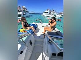 Two friends enjoy a sunny day on a boat, capturing memories while surrounded by a lively beach scene and fellow boaters in the turquoise waters.