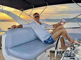 A man relaxes on the deck of a yacht at sunset, enjoying a drink and capturing the moment on his phone — a perfect scene for leisure and celebration on the water.