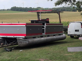 A sleek pontoon boat is docked on land, showcasing its modern design against a backdrop of lush fields and wind turbines.
