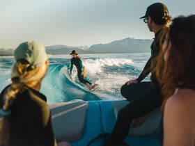 A thrilling moment captured as a surfer rides the wake behind a boat, with friends enjoying the scenic lake and mountain backdrop — perfect for adventure seekers.
