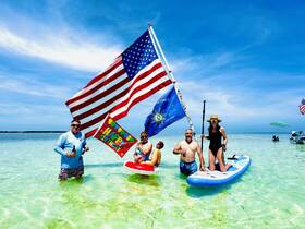 A lively group celebrates a birthday at a sandbar, showcasing festive flags and paddleboards in the clear waters — perfect for summer fun and memorable gatherings.