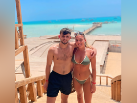 A happy couple poses together on a beach with a beautiful turquoise sea and wooden pier in the background, perfect for capturing summer memories.