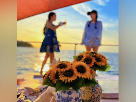 Two friends toast drinks against a stunning sunset backdrop on a boat, surrounded by vibrant sunflowers — a perfect moment for celebration and relaxation on the water.
