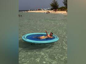 A man enjoys a sunny day lounging on a floating mat in clear waters, surrounded by beachgoers — a perfect scene for summer relaxation and fun.