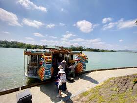 A colorful dragon boat awaits passengers at the riverbank, with guests preparing to board, showcasing a vibrant tour experience amidst scenic surroundings.