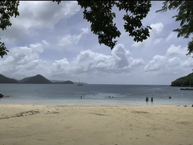A picturesque beach scene featuring people enjoying the water, with a sailboat in the distance and lush hills framing the coastline — perfect for a tropical getaway.