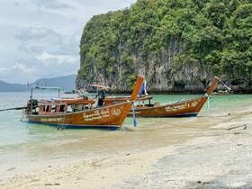Two traditional longtail boats are docked on a sandy beach, surrounded by lush greenery and rocky cliffs, showcasing a serene coastal scene.