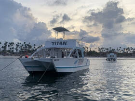 The 'Titan' catamaran is docked in calm waters, surrounded by a picturesque backdrop of palm trees and a beautiful sky, perfect for sunset cruises.