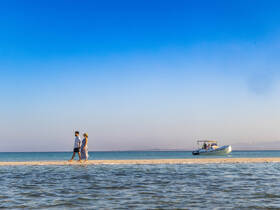 A couple strolls along a picturesque sandbar with a boat anchored nearby, capturing the essence of a perfect beach day.