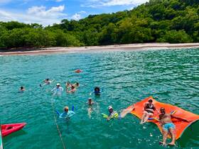 A lively group enjoys a sunny day swimming and relaxing on a floating mat in clear turquoise waters, perfect for a fun beach outing.