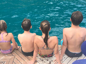 A group of children sits on the edge of a dock, eagerly looking into the clear blue water, ready for a fun day of swimming and adventure.