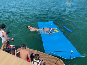 A lively group enjoys a fun day on the water, with kids playing on a floating mat and splashing around — perfect for summer adventures and family outings.