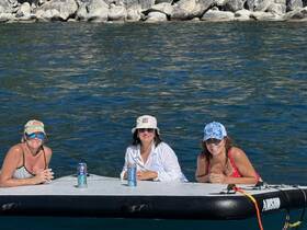 Three friends enjoy a sunny day on the water, relaxing on a paddleboard with drinks in hand — perfect for a fun day out with friends.
