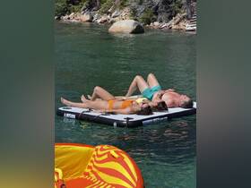 A group enjoys a relaxing day sunbathing on a floating mat in a serene lake setting, perfect for summer fun and water activities.