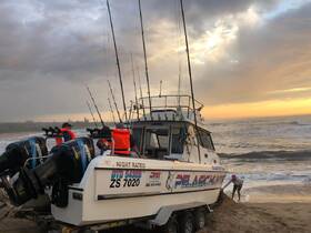 A group prepares for a fishing trip at sunset, with the boat 'Pelagic Hunter' beached on the shore — a perfect setting for an adventurous day on the water.