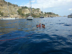 A group of friends enjoys a refreshing swim in clear waters, surrounded by sailboats and a picturesque coastline — perfect for a fun day on the water.