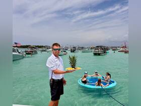 A lively scene featuring a server offering fresh pineapple to a group of friends enjoying a fun day on the water, surrounded by boats and clear turquoise waters.