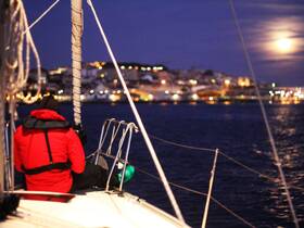 A sailor in a red jacket enjoys a serene night on the water, with the city lights twinkling in the background under the moonlight — a perfect moment for nighttime sailing adventures.