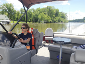 A young boy confidently steers a pontoon boat from the helm, showcasing the comfortable interior seating and a scenic waterway backdrop.