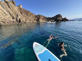Two individuals enjoy a refreshing swim in crystal-clear waters near rocky cliffs, with a boat anchored in the background — a perfect day for paddleboarding and exploring.