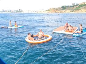 A lively group enjoys a day on the water, featuring paddleboarding and relaxing on a floating mat — perfect for summer fun and family outings.