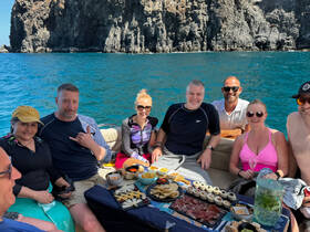 A cheerful group enjoys a boat party with a spread of delicious snacks and drinks, set against a stunning coastal backdrop — perfect for memorable outings on the water.