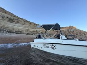 A side view of the 'Aegean Dream' boat anchored near a sandy beach, showcasing its sleek design against a backdrop of rocky cliffs.
