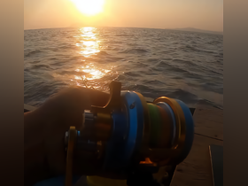 A hand holds a fishing reel against the backdrop of a stunning sunset over the ocean, capturing the essence of a peaceful fishing experience.