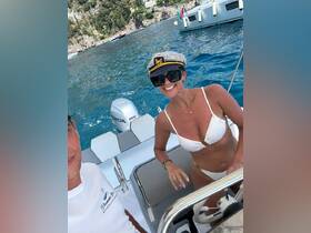 A joyful moment aboard a boat with a couple enjoying the sun, showcasing a fun day on the water with a captain's hat and beautiful scenery in the background.