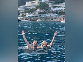 A joyful couple swims in the crystal-clear waters, waving at the camera with a stunning coastal backdrop — a perfect moment of fun and relaxation on their boating adventure.