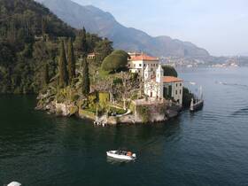 A picturesque view of a stunning lakeside villa surrounded by lush greenery and mountains, with a small boat navigating the serene waters — a perfect representation of scenic beauty and tranquility.