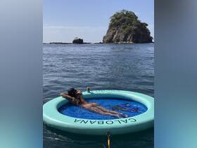 A woman relaxes on a floating mat in tranquil waters, with a picturesque island in the background — a perfect scene for a day of leisure on the water.
