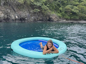 A joyful woman enjoys a relaxing moment on a floating mat in serene waters, surrounded by lush greenery — a perfect snapshot of fun and leisure on the water.