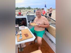 A man enjoys a slice of pizza on a boat, surrounded by friends at a beach — perfect for a fun day on the water.
