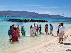 A joyful group celebrates a wedding on the beach, wading in the clear waters with stunning scenery in the background — a perfect setting for memorable moments.